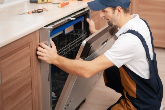 A man or service worker in special clothing installs, disassembles or performs maintenance of a dishwasher built into the kitchen furniture