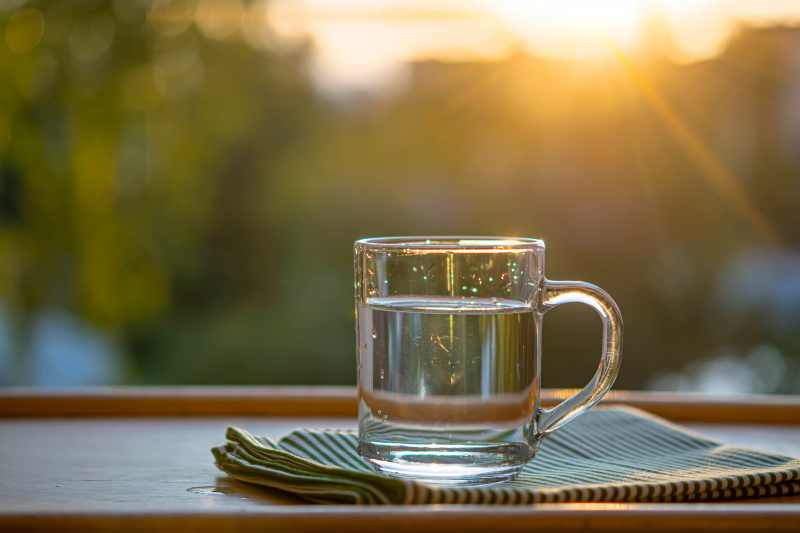 Clean drinking water in a glass cup with a handle stands on a napkin on a wooden table against the background of green nature.Sunset. The topic is thirst quenching, ecology. Selective focus
