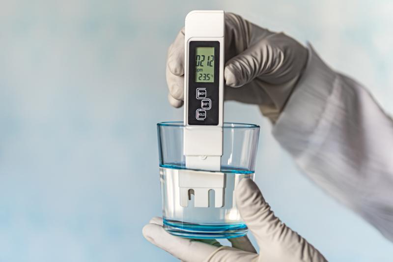 pH meter in hands with gloves, glass of water on blue background. Measurement of the characteristics of drinking water. The hardness of the water. poor water quality.