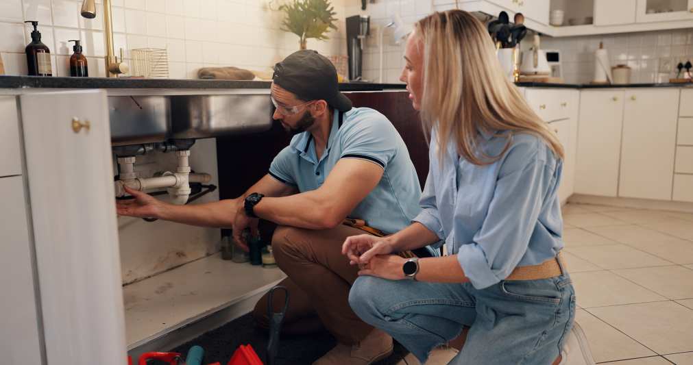 Plumber, man and woman by kitchen sink for pipe, inspection and check for leak for maintenance at house. People, handyman and client with assessment, repair and problem solving for drain at apartment.