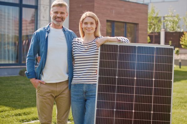 Happy married couple standing near solar panel. Man and woman cheerfully smiling with alternative energy source. Male and female with solar panel at lawn. Processing of solar energy into electricity.