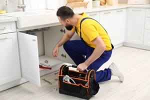plumber in uniform with toolbox fixing piping system in kitchen