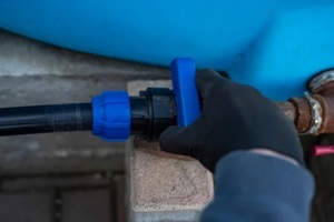 person in a black glove turning the blue handle of a plastic ball valve on a pipe connected to a blue water tank