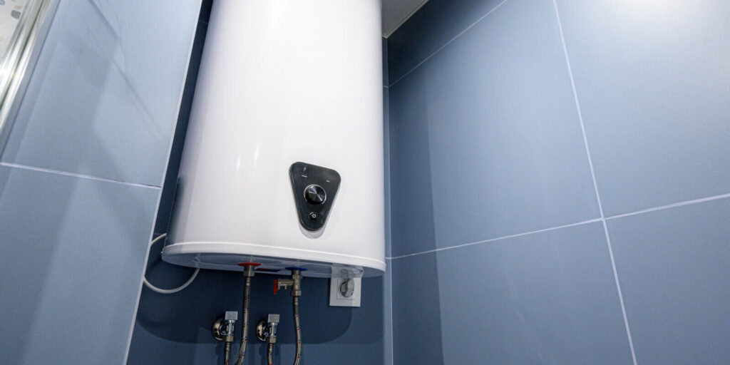 white electric water heater mounted on blue tiled wall in bathroom, with control knob, water inlet outlet pipes