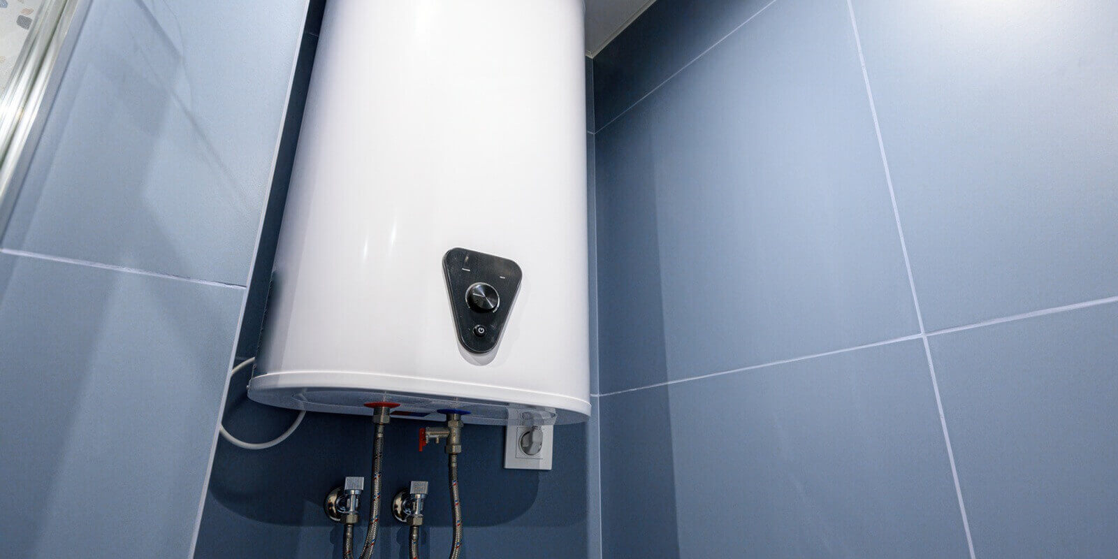 white electric water heater mounted on blue tiled wall in bathroom, with control knob, water inlet outlet pipes