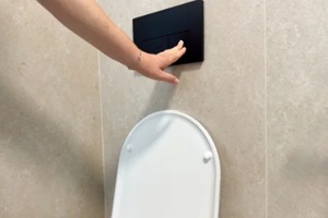 hand presses a black dual flush plate mounted on a beige tiled wall above an open toilet