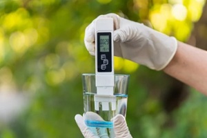 pH meter in hands with gloves, glass of water on blurred background of nature